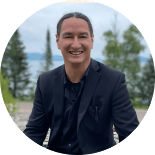 James-Vukelich-Kaagegaabaw-photo-yellowstone-forever A man with long dark hair in a low ponytail, wearing a black suit and shirt, smiles while seated outdoors with trees and water in the background.