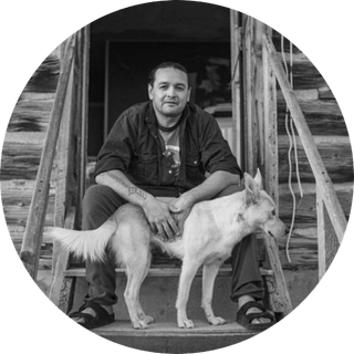 Jason-Baldes-photo-by-Russel-Albert-Daniels-yellowstone-forever-cultural-instructor A man sits on wooden steps outside a rustic cabin, looking at the camera, with a light-colored dog standing beside him.