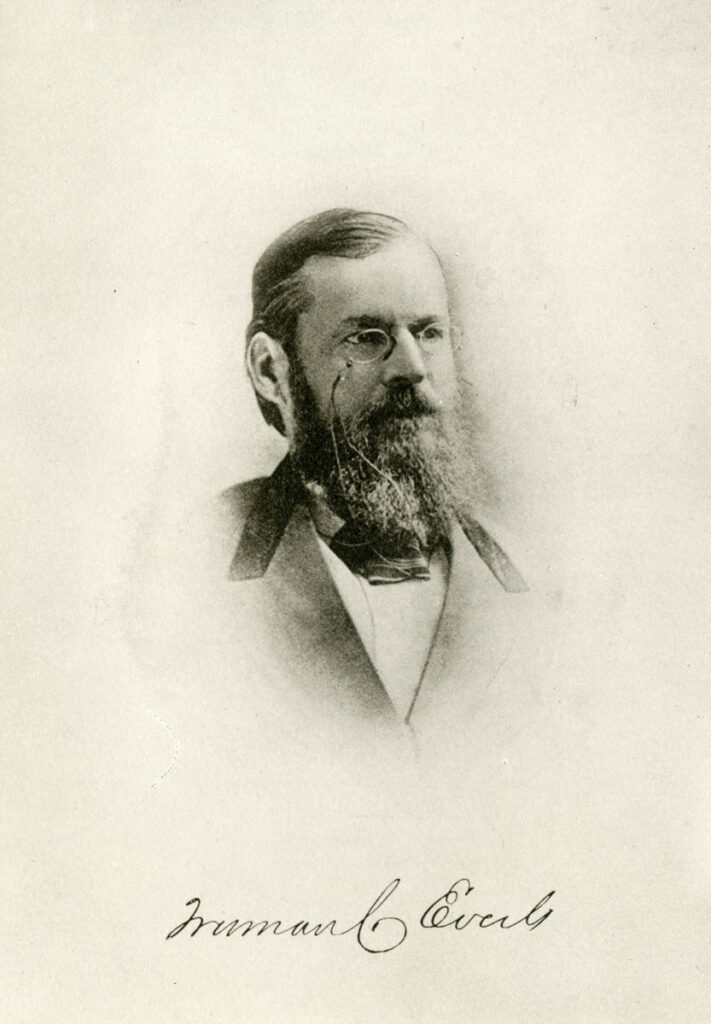 Black and white portrait of a bespectacled man with a full beard, wearing a suit and bow tie. His signature appears below the image.