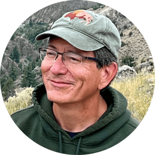 A man wearing glasses, a green hat with an animal logo, and a green hoodie, smiles outdoors with dry grassy hills and rocky terrain in the background.