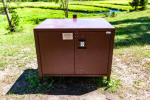 A brown bear-proof food storage locker stands on grass in a sunlit park, with trees, a stream, and greenery visible in the background.